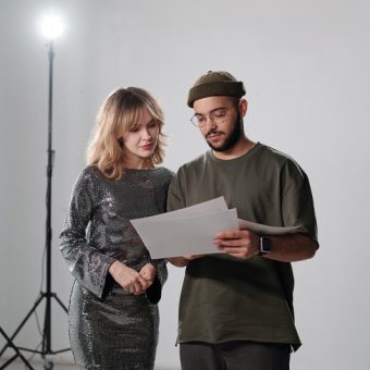 Vertical portrait of young actress consulting with assistant or director backstage and reading script notes in studio
